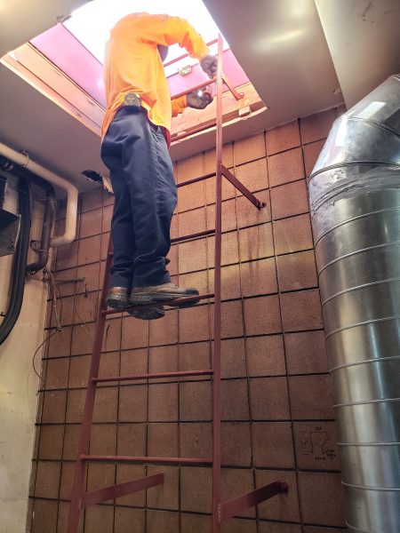 Inland Empire Fencing employee in orange shirt working on a ventilation repair at a commercial facility, highlighting specialized fencing and security solutions.