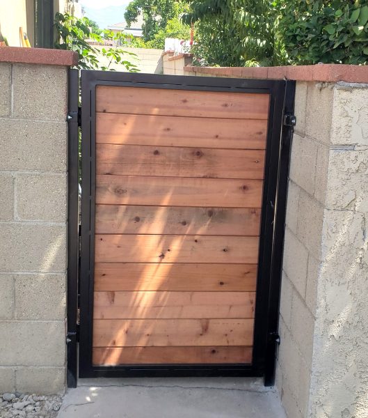 Sturdy single-panel wooden gate framed in black metal, set between cinder block walls, with a sunlit residential backdrop, showcasing Inland Empire Fencing’s durable and stylish entry solutions.